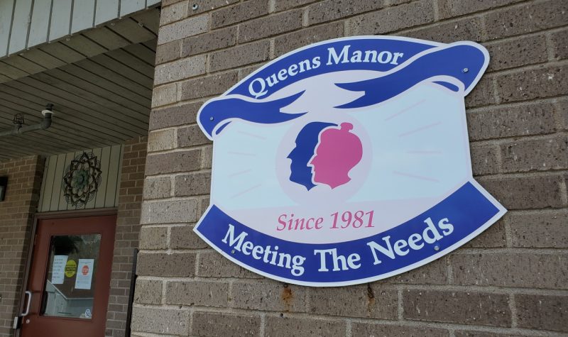 A blue, white and pink exterior sign for Queens Manor. It has two outlines of heads on it, one male and one female, in pink and blue.