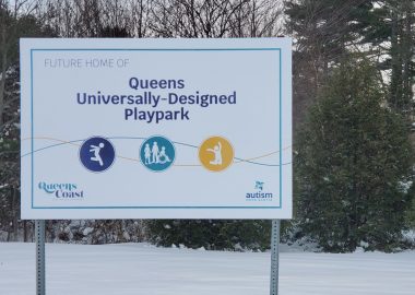 A sign in front of a snowy stand of trees indicates a playground is to be built in that location