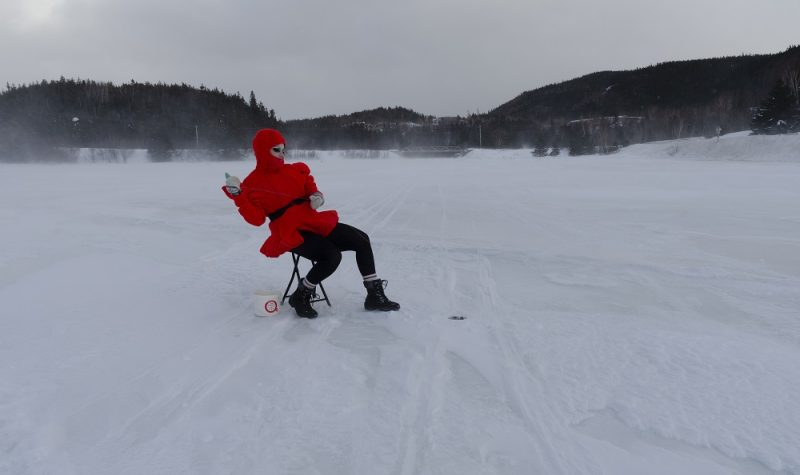 The Queer Mummer sits in winter clothing on a folding chair on a frozen body of water.