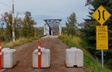 Cement blocks and orange cones are lined in front of a bridge in the distance. There is forest on either side and it is a sunny day.