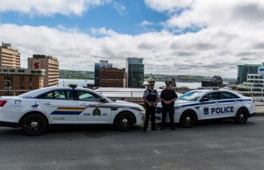 Une voiture de la GRC fait face à une voiture de la police d'Halifax. Les deux officier pose devant leur véhicule. Ils sont devant un paysage d'Halifax, on voit les building et le port au fond.