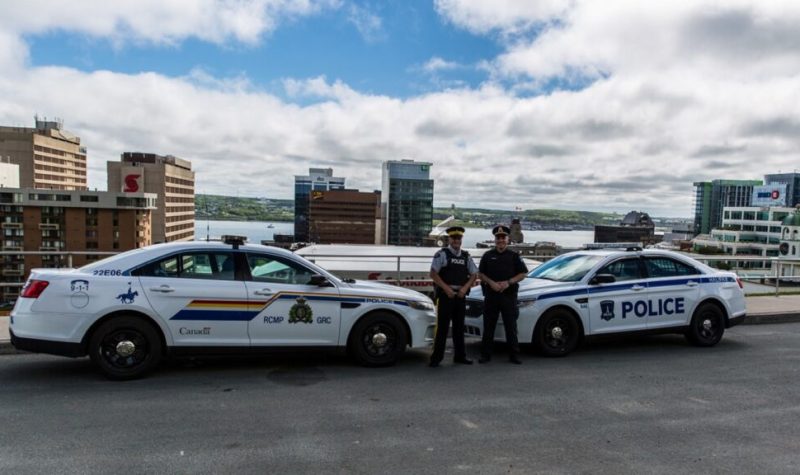 Une voiture de la GRC fait face à une voiture de la police d'Halifax. Les deux officier pose devant leur véhicule. Ils sont devant un paysage d'Halifax, on voit les building et le port au fond.