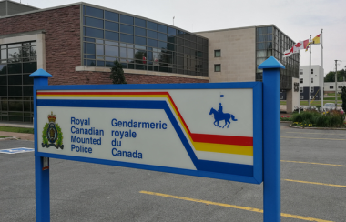 A blue, yellow, red and white sign with the crest and name of the Royal Canadian Mounted Police. The sign is in the foreground of the photo next to a parking lot and in front of a two story brick building with grey glass windows.