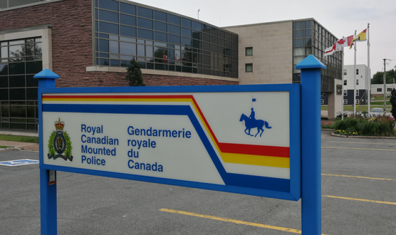 A blue, yellow, red and white sign with the crest and name of the Royal Canadian Mounted Police. The sign is in the foreground of the photo next to a parking lot and in front of a two story brick building with grey glass windows.