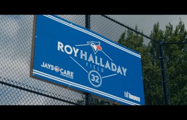 A blue sign with white text on a fence outdoors. A green tree is in the distance.