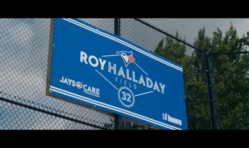 A blue sign with white text on a fence outdoors. A green tree is in the distance.