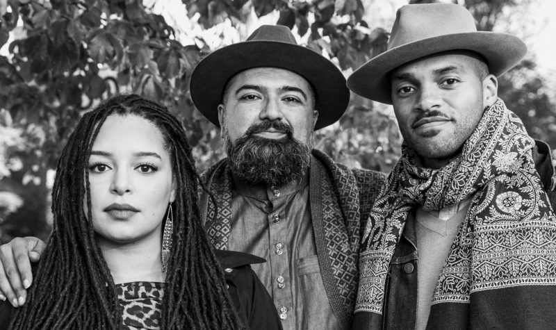 A black and white photo of three musicians standing in front of a tree. Ruby Singh stands in the middle of his band members The Future Ancestors.