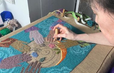 Dame travaillant sur un tapis en laine représentant des mains disposées en cercle.