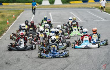 A number of kids drive racing karts into a corner, the lead kart is dark blue and there is a lot of drivers wearing red racing suits. In the background two track employees hold safety flags for any emergencies.