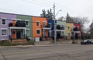 A row of seven brightly coloured townhouses next to a paved street.