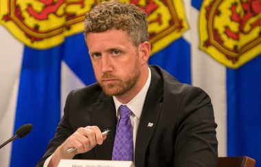 Nova Scotia Premier Iain Rankinn sits at a podium at a press conference with flags behind him.