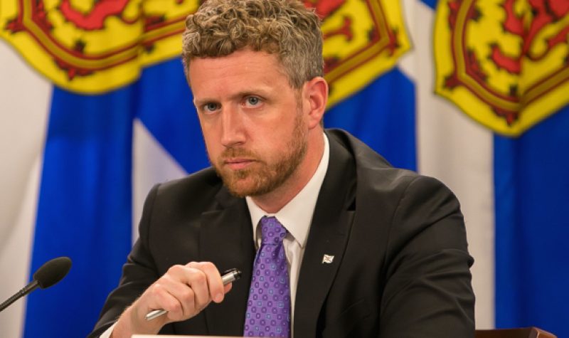 Nova Scotia Premier Iain Rankinn sits at a podium at a press conference with flags behind him.