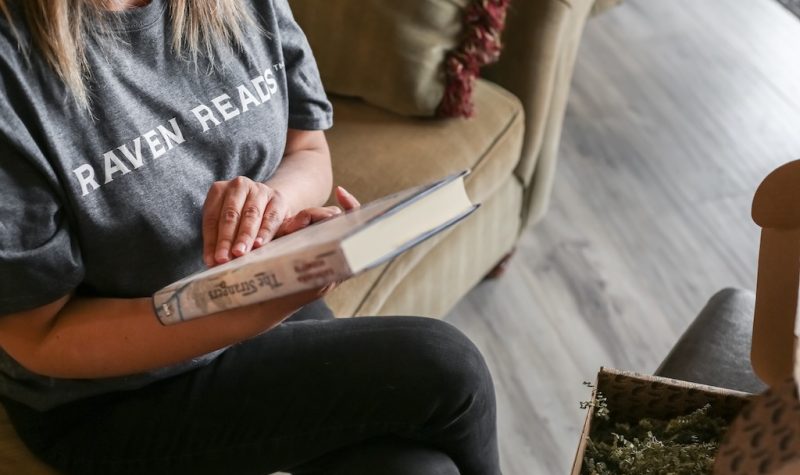 A woman reading a book with a shirt that says 'Raven Reads'