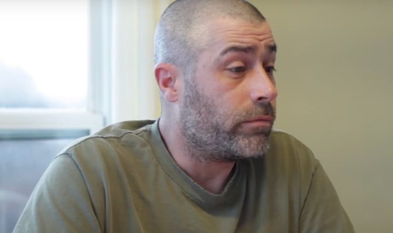 A man with close-cropped hair and a light beard wearing an olive green T-shirt sits by a window.