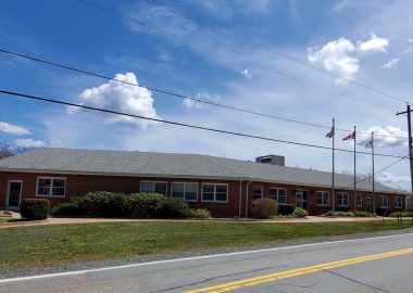 A long, brick building is seen next to a road on a sunny day with telephone lines above