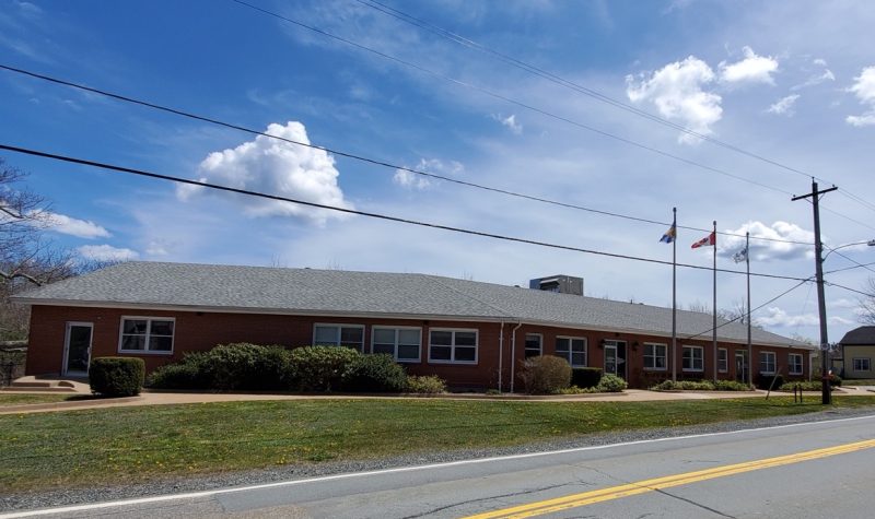 A long, brick building is seen next to a road on a sunny day with telephone lines above.