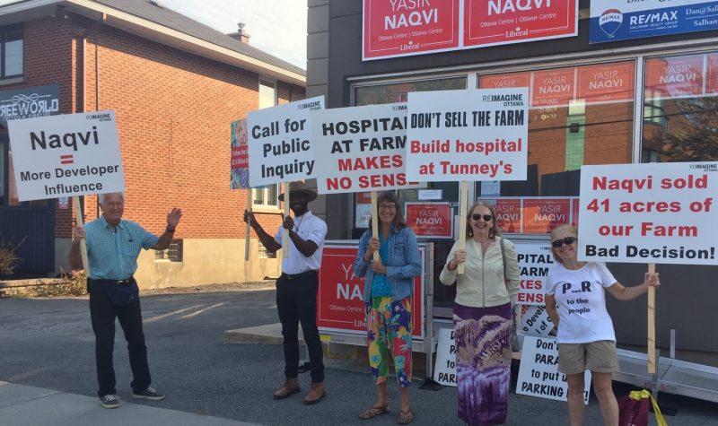 On a sunny day, a group of protestors pickets on a curb outside of MP Yasir Naqvi's office.