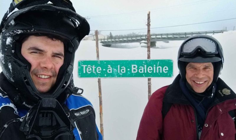 Deux hommes portant un casque de motoneige font face à la caméra devant un décor enneigé. Entre eux, on peut lire un panneau de signalisation indiquant Tête-à-la-Baleine.