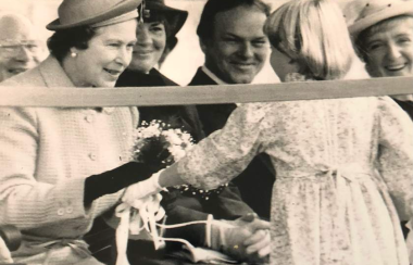 A black and white photo of a small girl handing Queen Elizabeth flowers with other adults looking on.