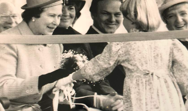 A black and white photo of a small girl handing Queen Elizabeth flowers with other adults looking on.