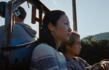 Two people siting down outside look to the right of them. The sky is blue with trees around them.