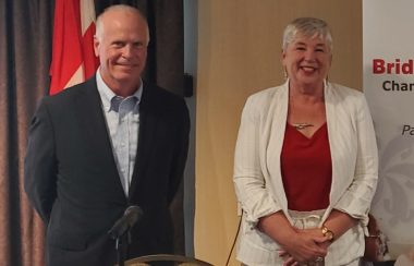 A male and female politician stand in front of a table with a microphone with a Canada flag behind them
