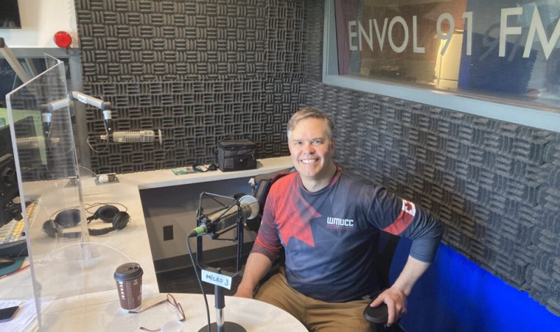 Une photo de Robert Loiselle, président du comité d'organisation de Chef en plein air. Un homme d'âge moyen aux cheveux grisonnants, assis sur une chaise dans le studio de radio d'Envol 91 FM. il porte un haut de sport bleu et rouge et un short beige et fait un grand sourire à la caméra.