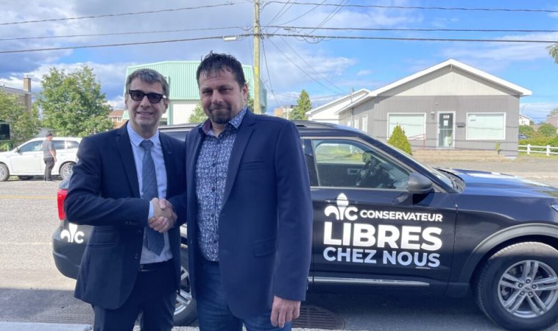Deux hommes se serrant la main devant un véhicule sur lequel on peut voir le logo du Parti conservateur du Québec avec l'inscription : Libres chez nous.