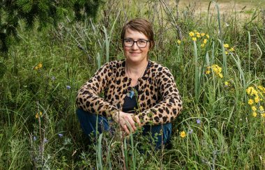 A woman with light brown hair, wearing glasses and a patterned shirt sits in a field of tall grass and flowers.