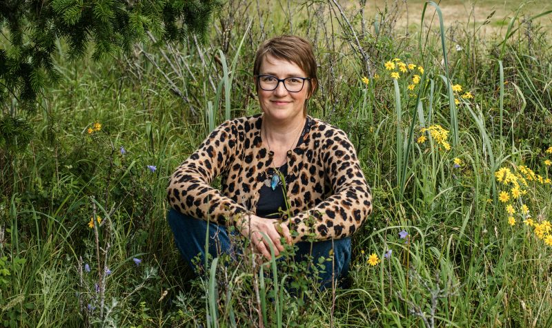 A woman with light brown hair, wearing glasses and a patterned shirt sits in a field of tall grass and flowers.