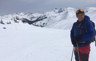 A body shot of Ron dart with ski poles in his hand surrounded by mountains.