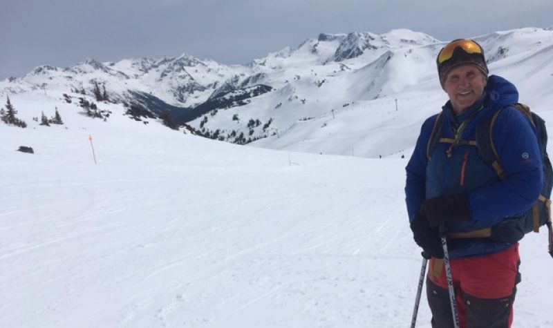 A body shot of Ron dart with ski poles in his hand surrounded by mountains.