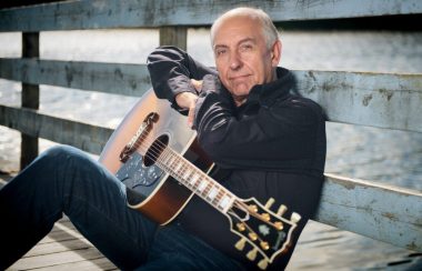 Homme avec chemise et culottes noires assis avec une guitare sur un banc.