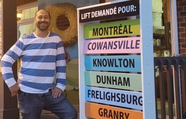 Pictured is Frédéric Gauld standing outside the bagel shop with his transportation panel. Places such as Montreal, Cowasnville, Knowlton, Dunham, etc., are listed in bright colours.