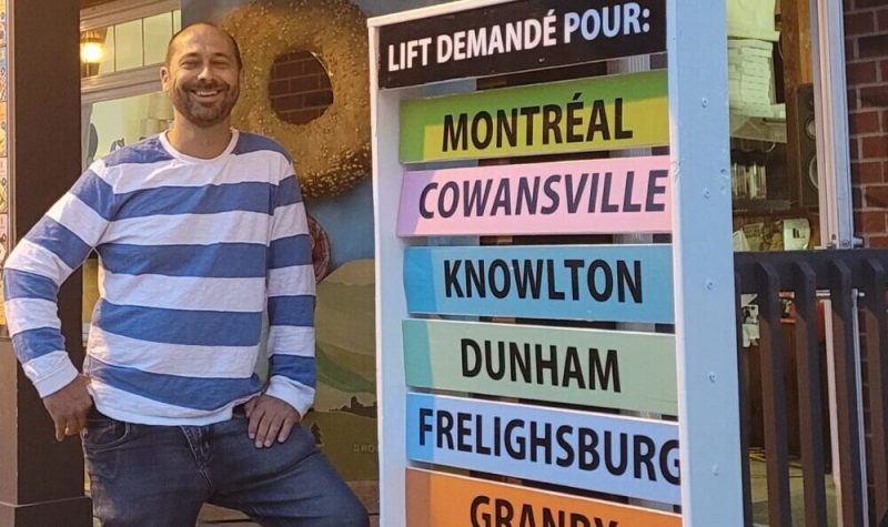 Pictured is Frédéric Gauld standing outside the bagel shop with his transportation panel. Places such as Montreal, Cowasnville, Knowlton, Dunham, etc., are listed in bright colours.