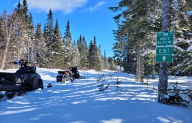 Deux motoneiges sur un sentier enneigé se trouvant au centre de rangées de conifères. À droite du sentier, un panneau indiquant la Route blanche direction Est.