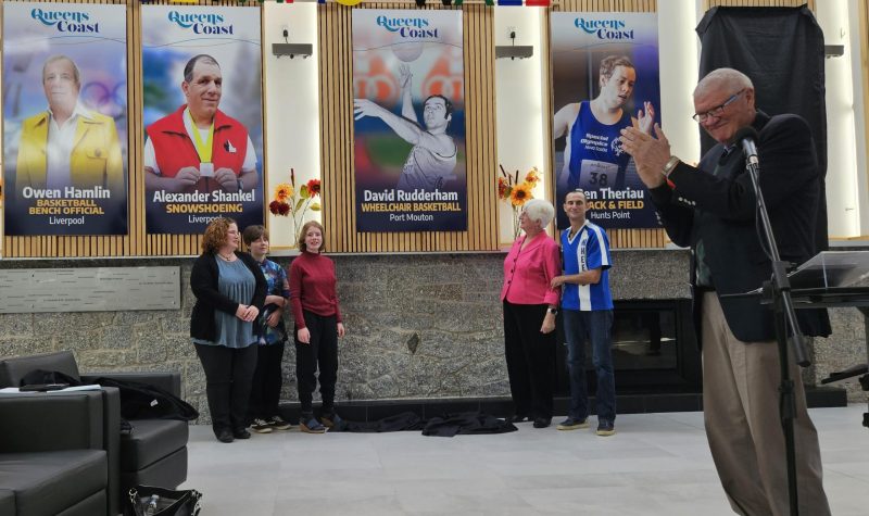 A man applauds in the foreground as a family stands beneath a portrait of an Olympic wheelchair basketball athlete. The portrait is unveiled on a wall above their heads.