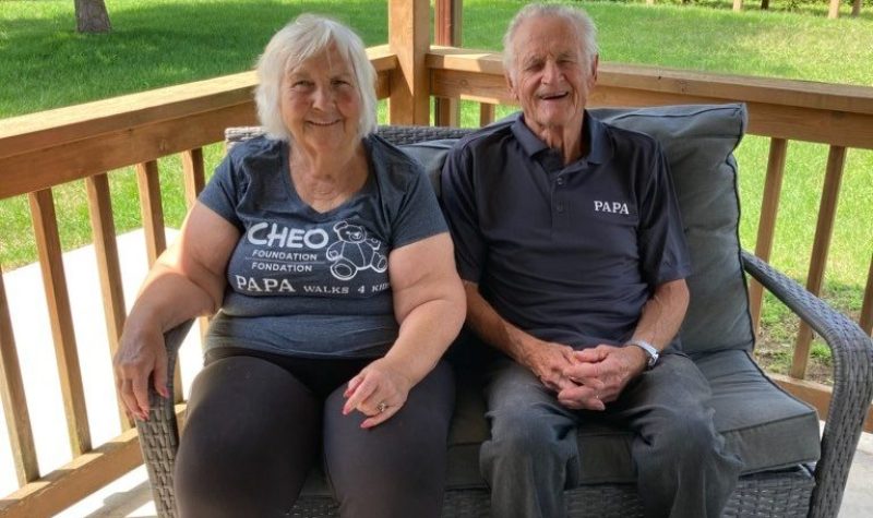 An elderly woman and man sit on a couch on a porch.