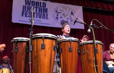 A man on a stage plays a set of five tall hand drums.