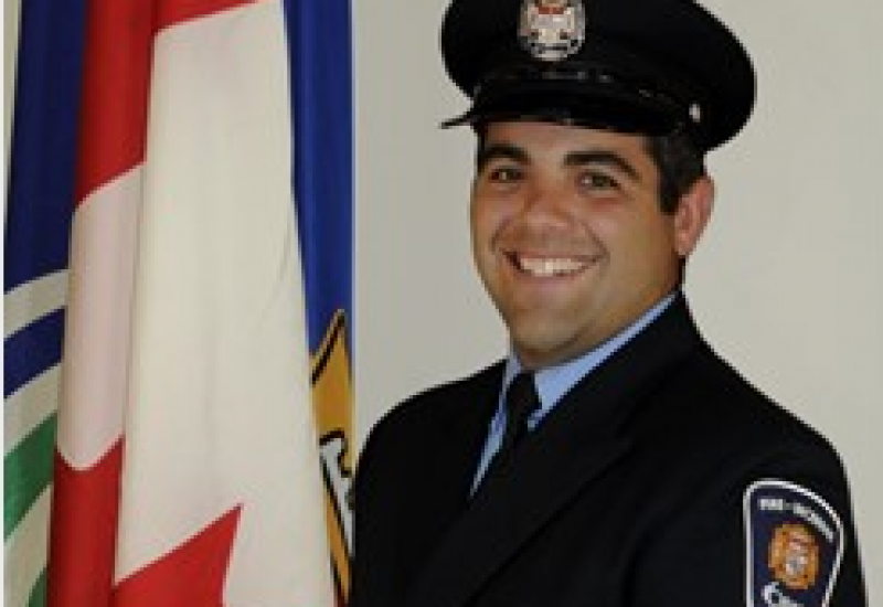 A man wearing a formal black uniform stands slightly turned, smiling at the camera, the Ottawa and Canada flags to his left.