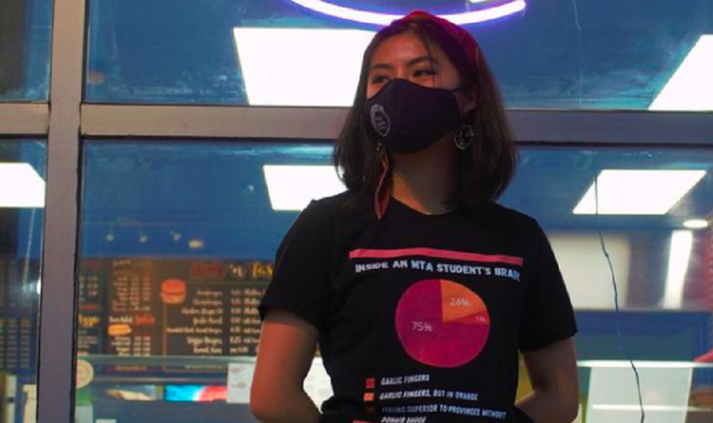 A person wears a T-shirt and a face mask while standing in front of a restaurant.