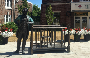 A bronze sculpture of Sir John A. Macdonald alongside a bronze stylized prisoners' box with potted flowers in the background. Statue is situated in front of Picton Armoury.