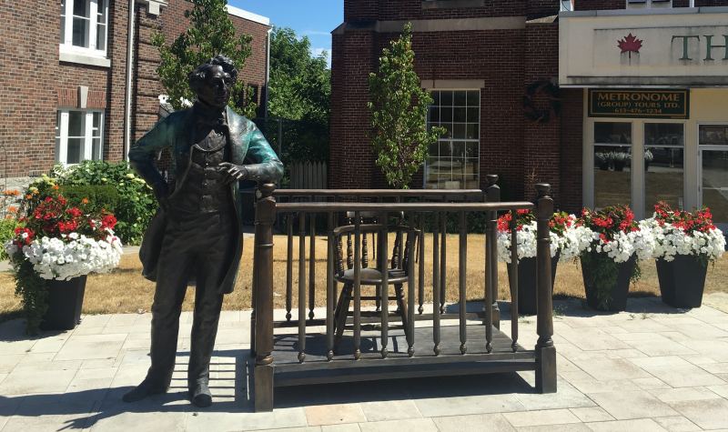A bronze sculpture of Sir John A. Macdonald alongside a bronze stylized prisoners' box with potted flowers in the background. Statue is situated in front of Picton Armoury.