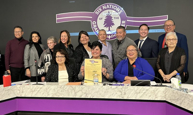 Thirteen people behind a table against a dark grey wall containing a purple logo saying Six Nations Grand River