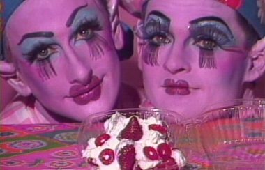 Two people in drag makeup rest their chins on a table with whipped cream and strawberries in a container in front of them.