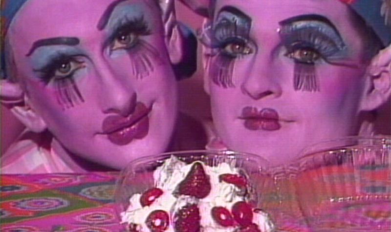 Two people in drag makeup rest their chins on a table with whipped cream and strawberries in a container in front of them.