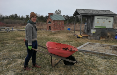 A woman holding the handles of a wheelbarrow in a spring garden area with a composter, tool shed, and brick oven behind her.