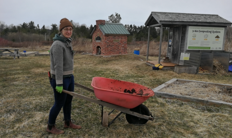 A woman holding the handles of a wheelbarrow in a spring garden area with a composter, tool shed, and brick oven behind her.
