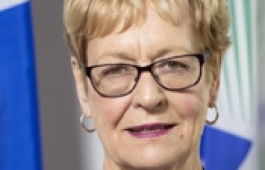 A professional headshot of Shawville Mayor Sandra Murray, wearing a purple shirt and glasses, standing in front of a Quebec flag.