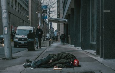 Une personne dort sur le trottoir d'une grande ville. Les passants y sont indifférents.